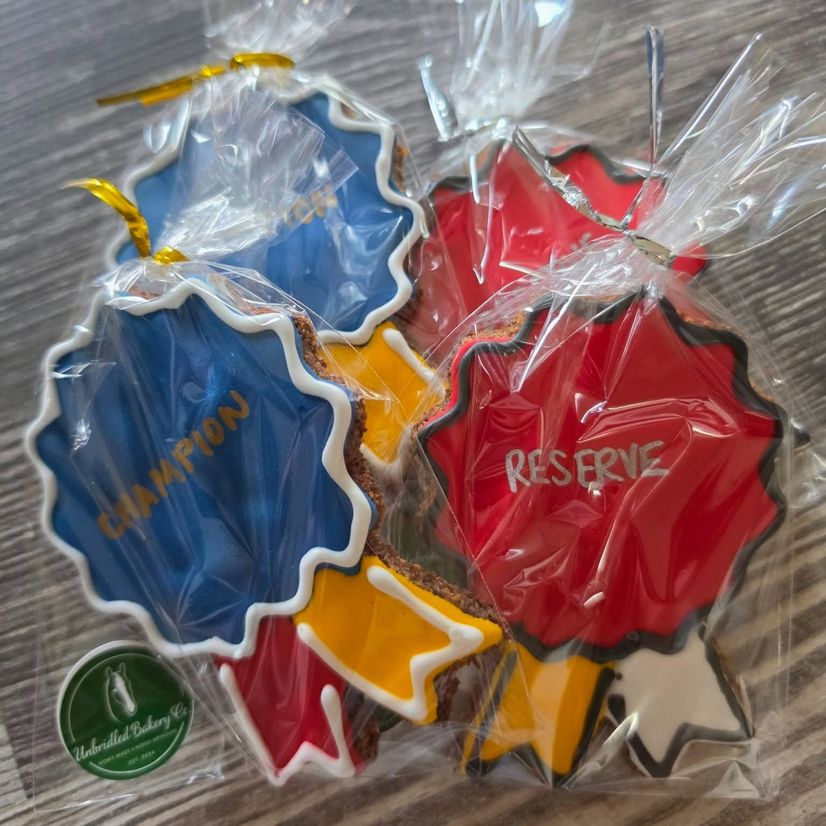 Decorated horse cookies shaped like  championship and reserve champion ribbons, packaged in clear cellophane bags on a gray wood surface. One ornament is blue with gold trim and the word 'CHAMPION', one is red with white trim and the word 'RESERVE', and one is yellow with black trim.