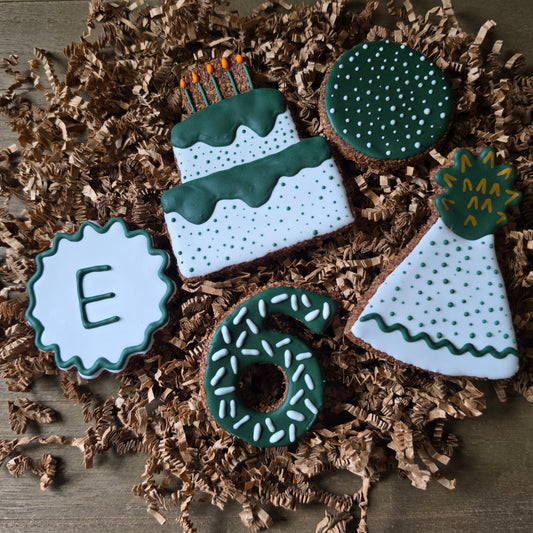 Decorated apple flax horse cookies arranged on brown shredded paper over a dark wood surface. The cookies include a round cookie with the letter E, a tiered birthday cake, a green dotted circle cookie, a party hat, and a number 6 cookie, all in white and dark green icing with small orange candle accents.