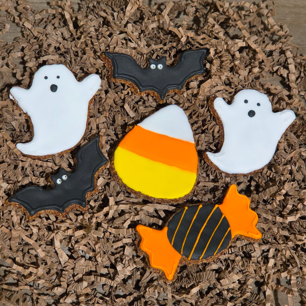 Halloween-themed apple flax horse cookies, including two white ghosts, two black bats, a candy corn cookie, and an orange and black striped candy cookie, arranged on brown shredded paper over a wooden surface.