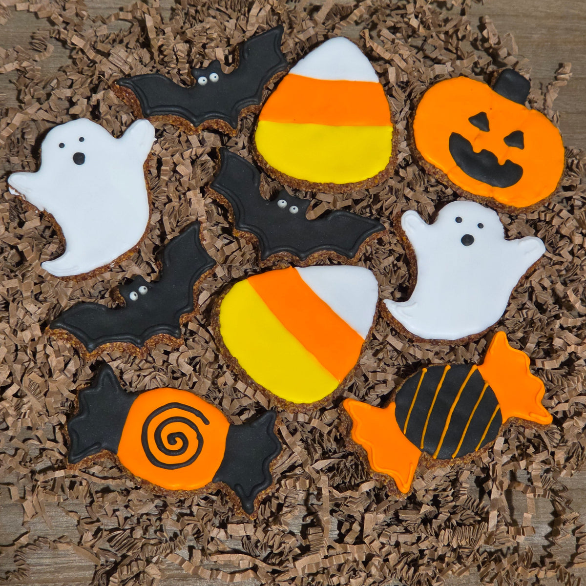 A set of Halloween-themed apple flax horse cookies decorated with white ghosts, black bats, orange pumpkins or jack-o-lanterns, candy corn, and an orange-and-black striped candy, arranged on brown shredded paper over a wooden surface.