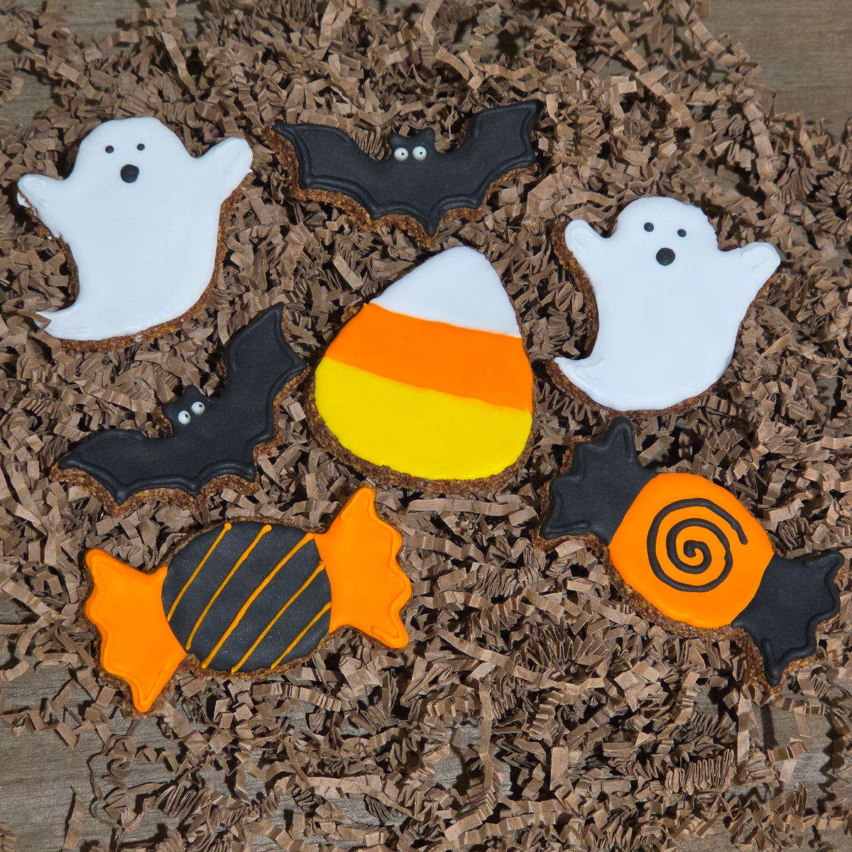Halloween-themed apple flax horse cookies decorated as ghosts, bats, candy corn, and wrapped candy arranged on brown shredded paper over a wooden surface.