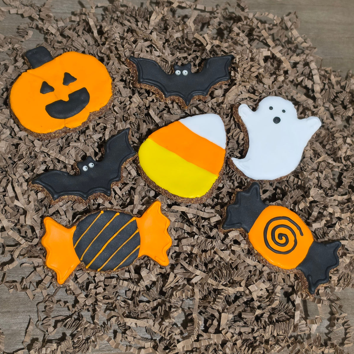 Halloween-themed apple flax horse treats decorated with orange, black, white, and yellow icing, arranged on brown shredded paper over a wooden surface.