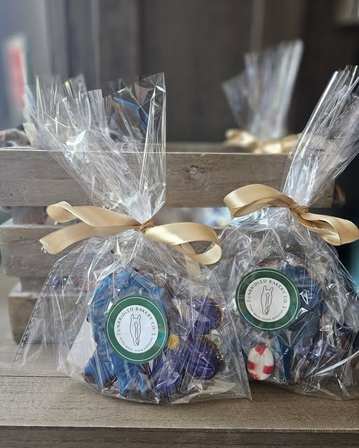 Two clear cellophane gift bags tied with beige ribbons, filled with blue and purple wrapped horse cookies displayed on a wooden surface. Each bag has a round green-and-white label reading Unbridled Bakery Co.